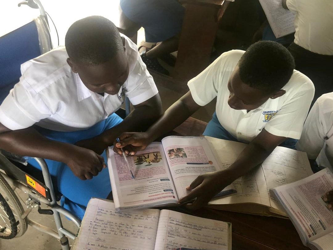 A girl in a wheelchair is sitting next to a classmate at a desk. They are looking at a textbook together