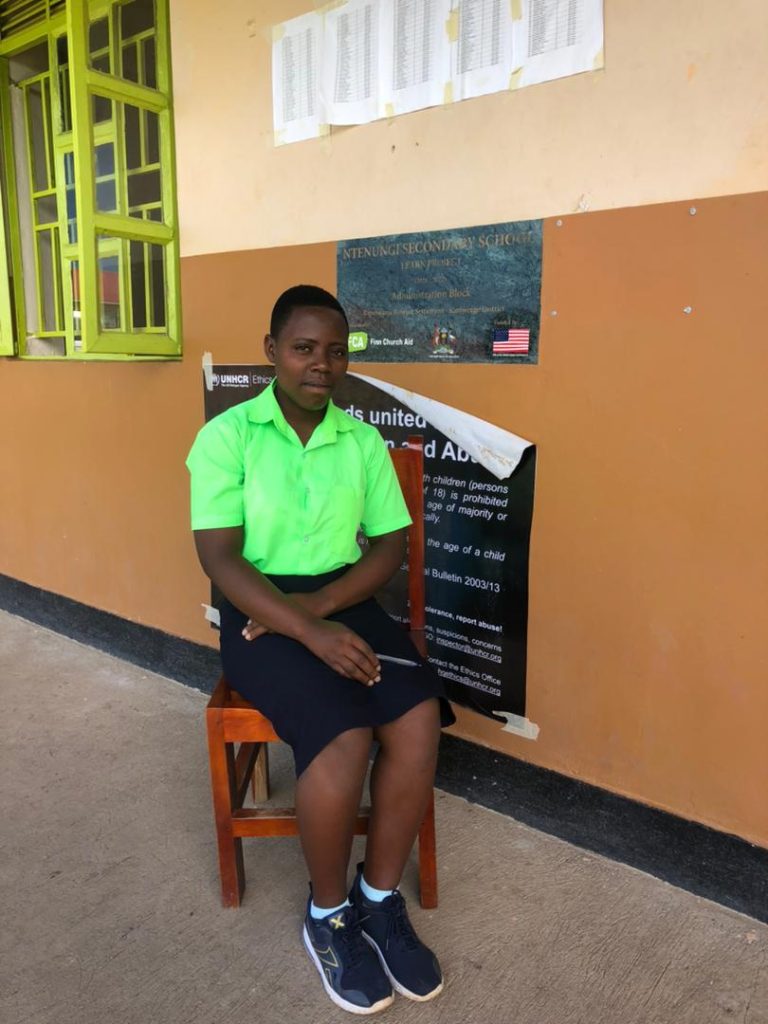 A girl in a green polo shirt sits on a chair outside a classroom and looks towards the camera