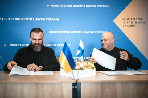 Two men sit at a table signing documents. A Ukrainian and Finnish flag is on the table next to some flowers.