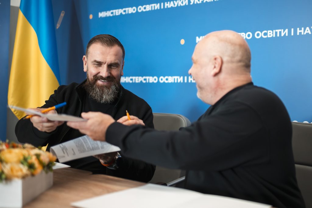 Kaksi miestä istuu pöydän ääressä. Toinen heistä antaa papereita toiselle ja kumpikin hymyilee. Taustalla näkyy Ukrainan lippu.