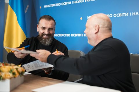 Two men sit at a table exchanging documents while smiling. A Ukrainian and Finnish flag is on the table next to some flowers.