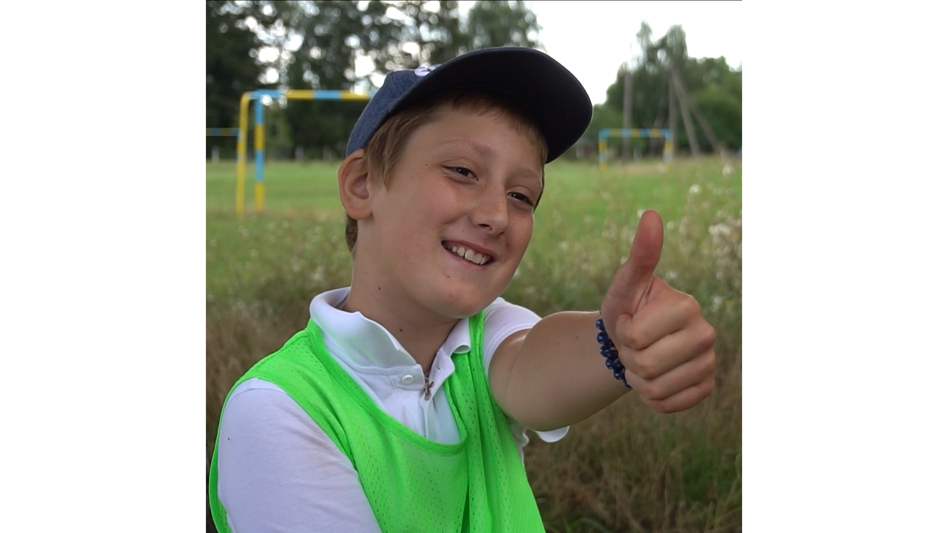 A boy in a baseball cap and a green vest smiles and shows the thumbs up sign