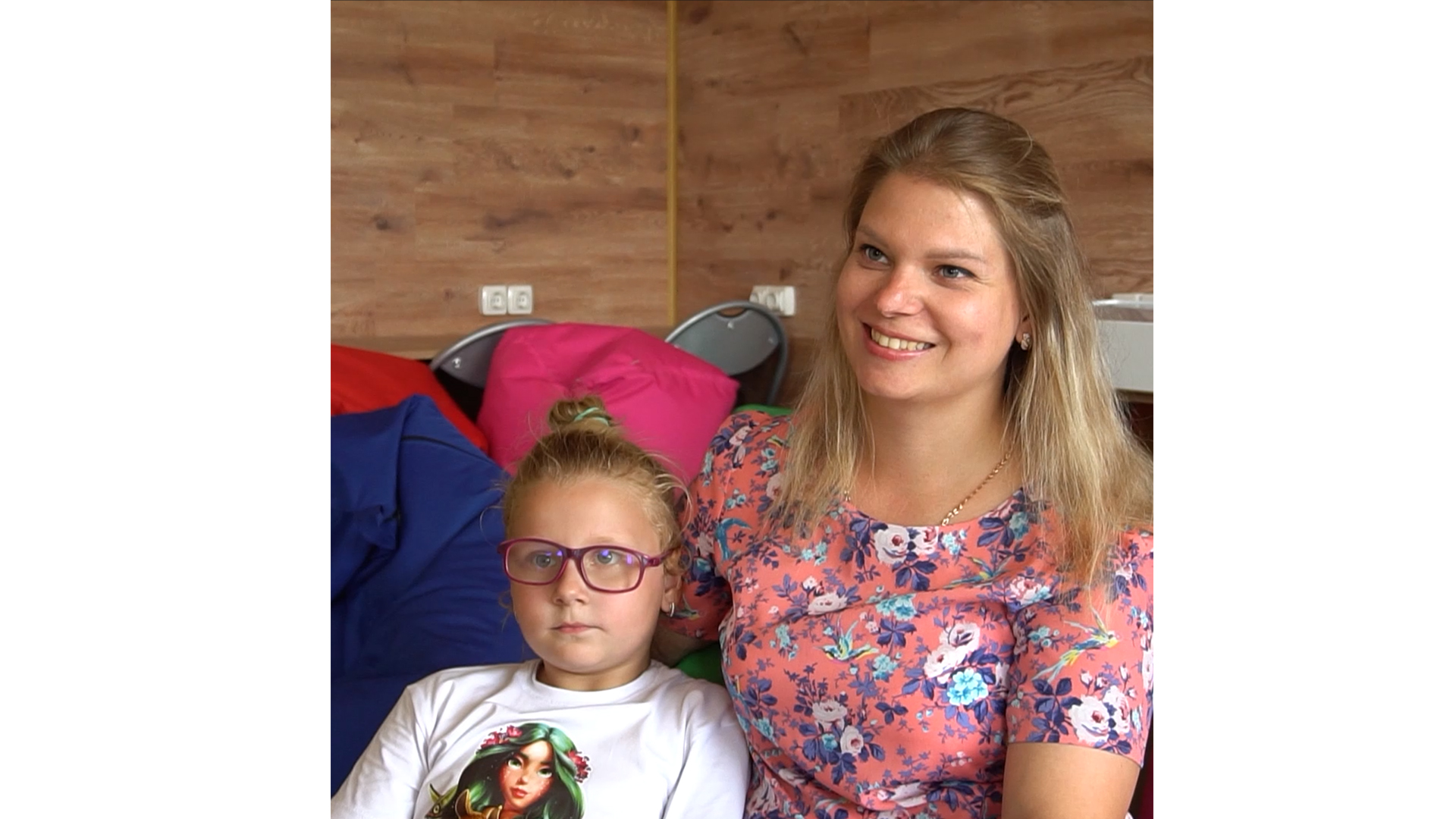 A smiling woman in a pink flowery top sits in a room with many multi-coloured beanbags. A child sits closely next to her.