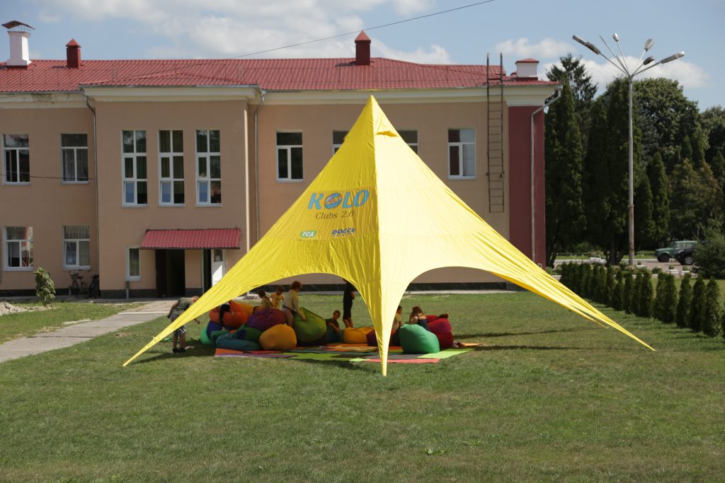 A yellow canopy in front of a school. Beanbags lie under the canopy. 