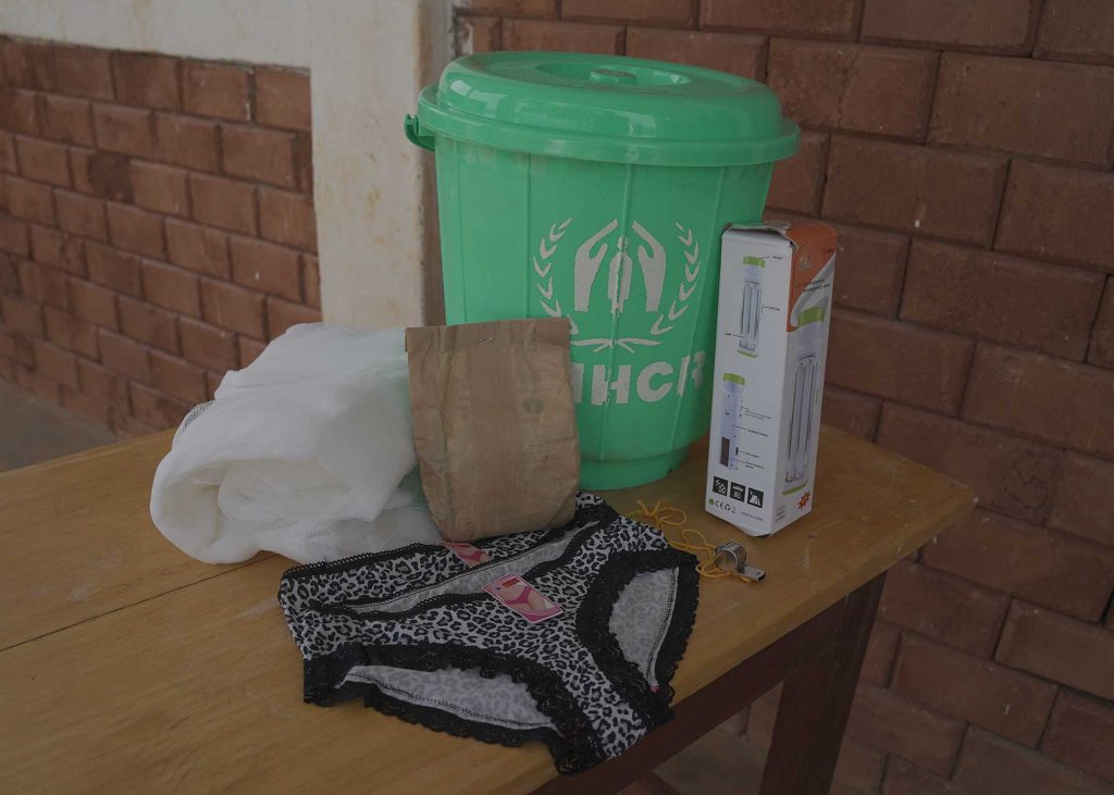 Some materials from FCA's and UNHCR's dignity kit in are spread on the table. There is a bucket, a box with a picture of a torch, a whistle, a paper bag, two pairs of women's underwear and a white mosquito net on the table. 