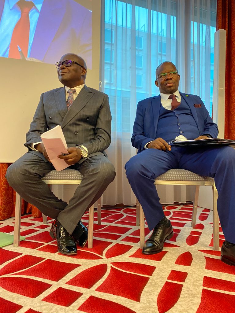 Two men in suits sit on chairs on a brightly patterned carpet