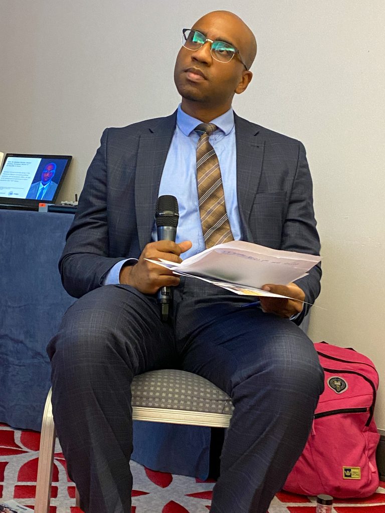 A man in a suit sits on a chair and listens. He has a microphone in his hand