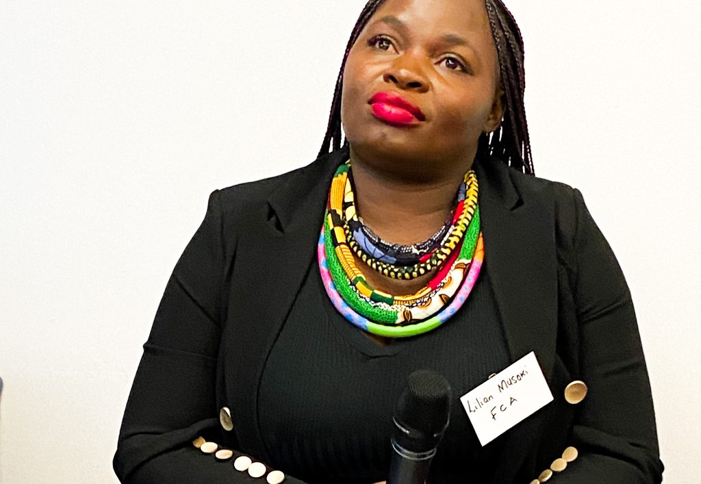 A woman in a black suit wearing a bright necklace and red lipstick listens intently.