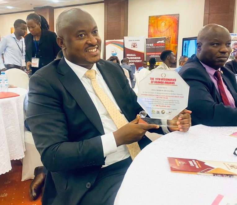 A smiling man in a suit sits at a table in a room full of people. He is holding an award and showing it to the camera.