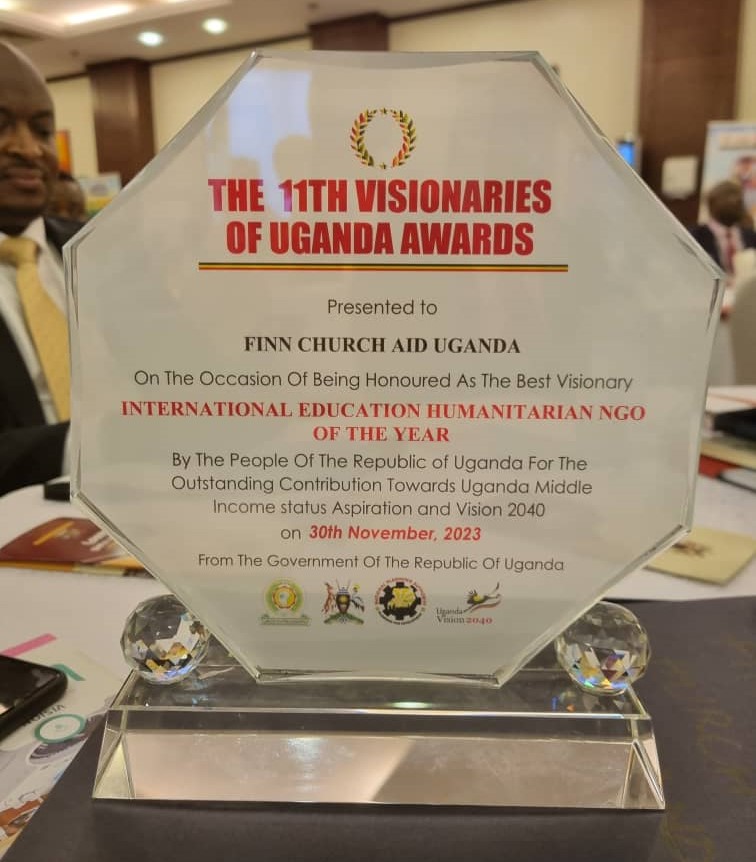A large glass award. Text engraved on it reads "THE 11TH VISIONARIES OF UGANDA AWARDS Presented to FINN CHURCH AID UGANDA On The Occasion Of Being Honoured As The Best Visionary INTERNATIONAL EDUCATION HUMANITARIAN NGO OF THE YEAR By The People Of The Republic of Uganda For The Outstanding Contribution Towards Uganda Middle Income status Aspiration and Vision 2040 on 30th November, 2023 From The Government Of The Republic Of Uganda"
