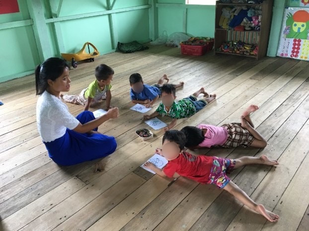 A woman sits on a wooden floor and talks to five children who are lying on the floor