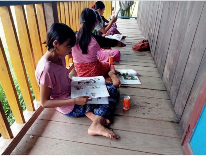 A row of girls sit on a wooden balcony reading books