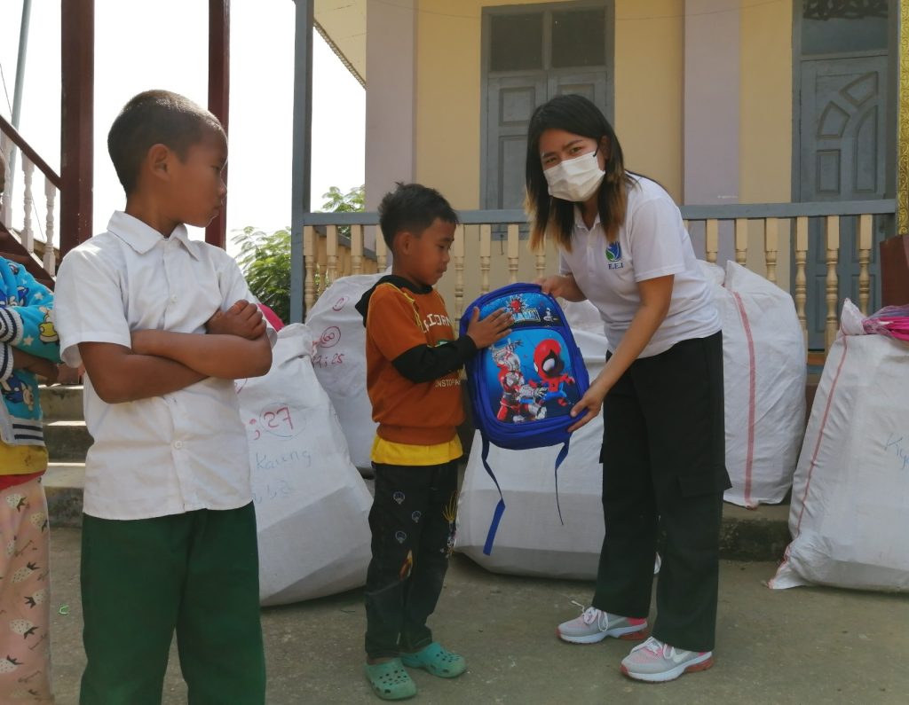 A woman in a mask gives a backpack to a child outside a school while another child looks on.