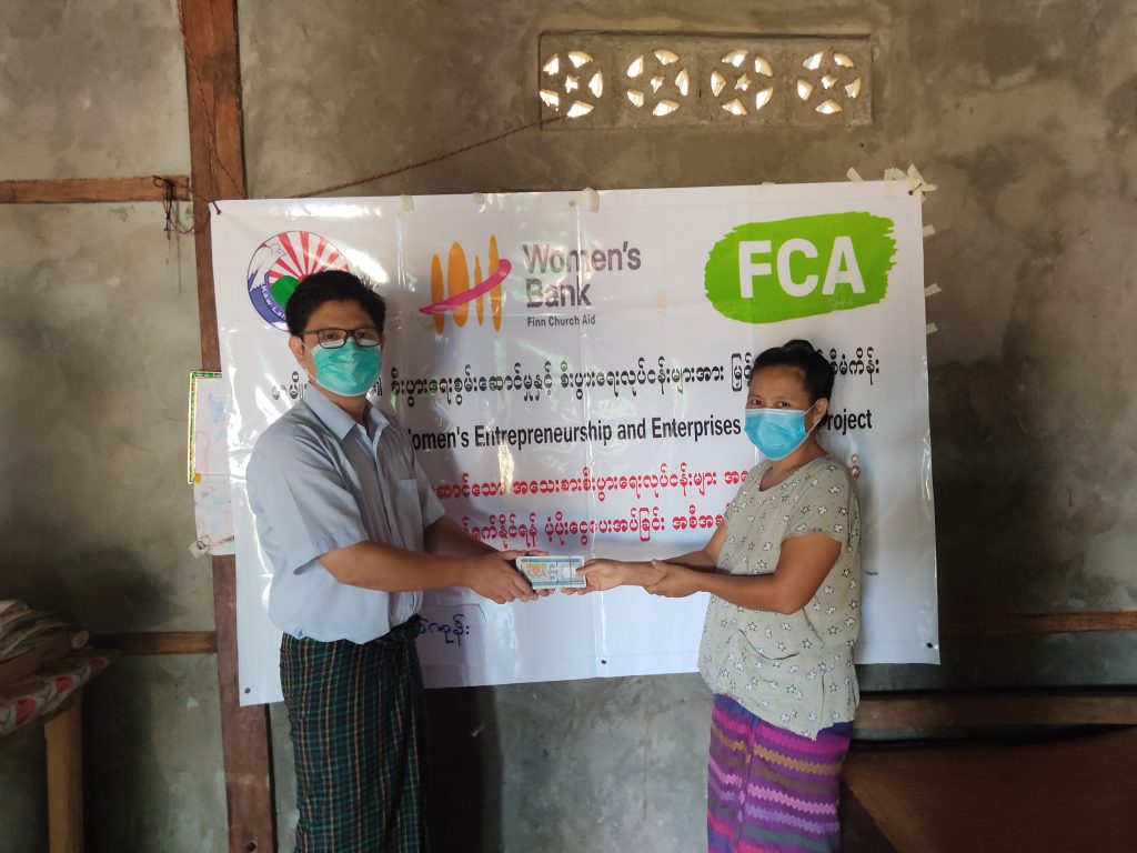 A man and a woman in face masks pose for the camera in a room with concrete walls. The man is handing money to the woman. On the wall hangs a banner with writing in Burmese and the logos of the Kaw Lah Foundation, Women's Bank and FCA