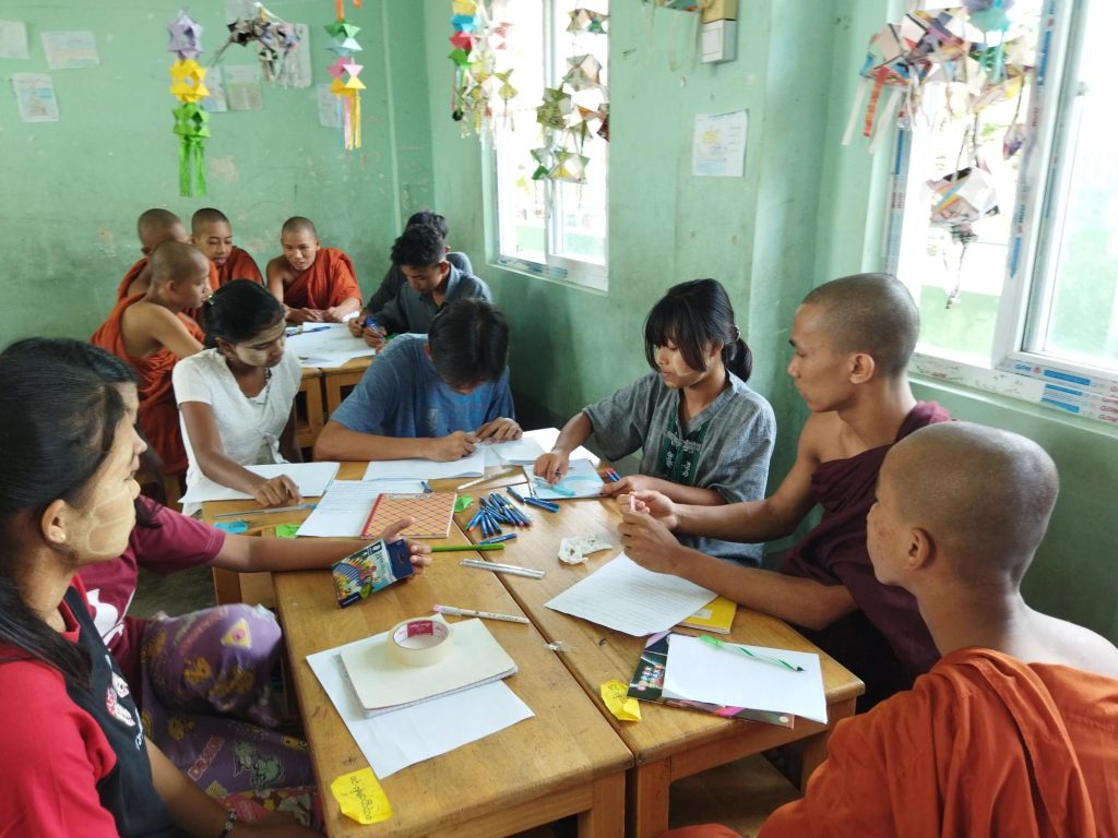 A number of teenage students, some in buddhist robes, sit in a classroom at desks writing with great concentration