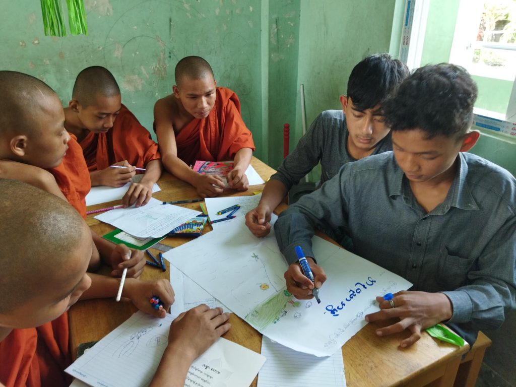 A group of boys, some in Buddhist robes sit around a desk and write on paper