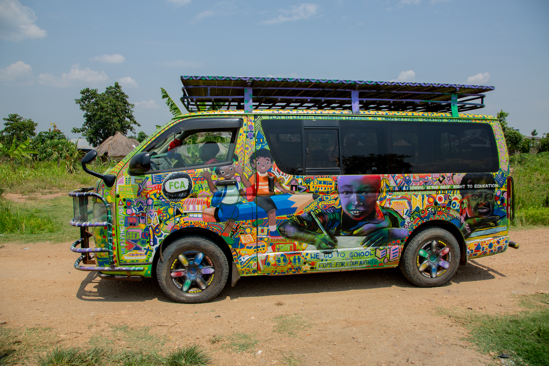 A brightly coloured minibus with numerous illustrations of children, positive slogans and the FCA logo stands on a dirt road.