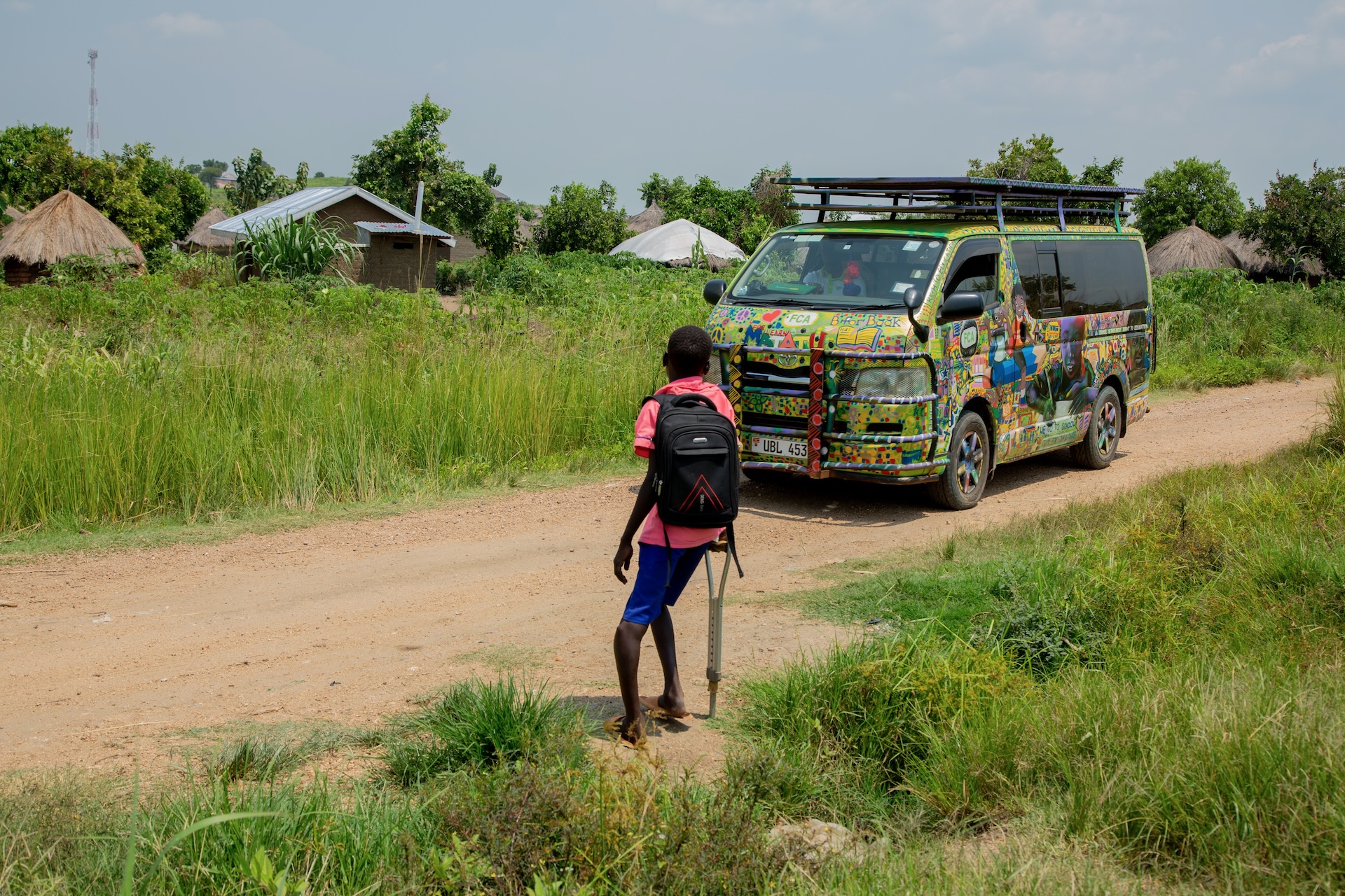 A schoolchild wearing a rucksack and leaning on a crutch waits for a brightly coloured bus on a dirt road
