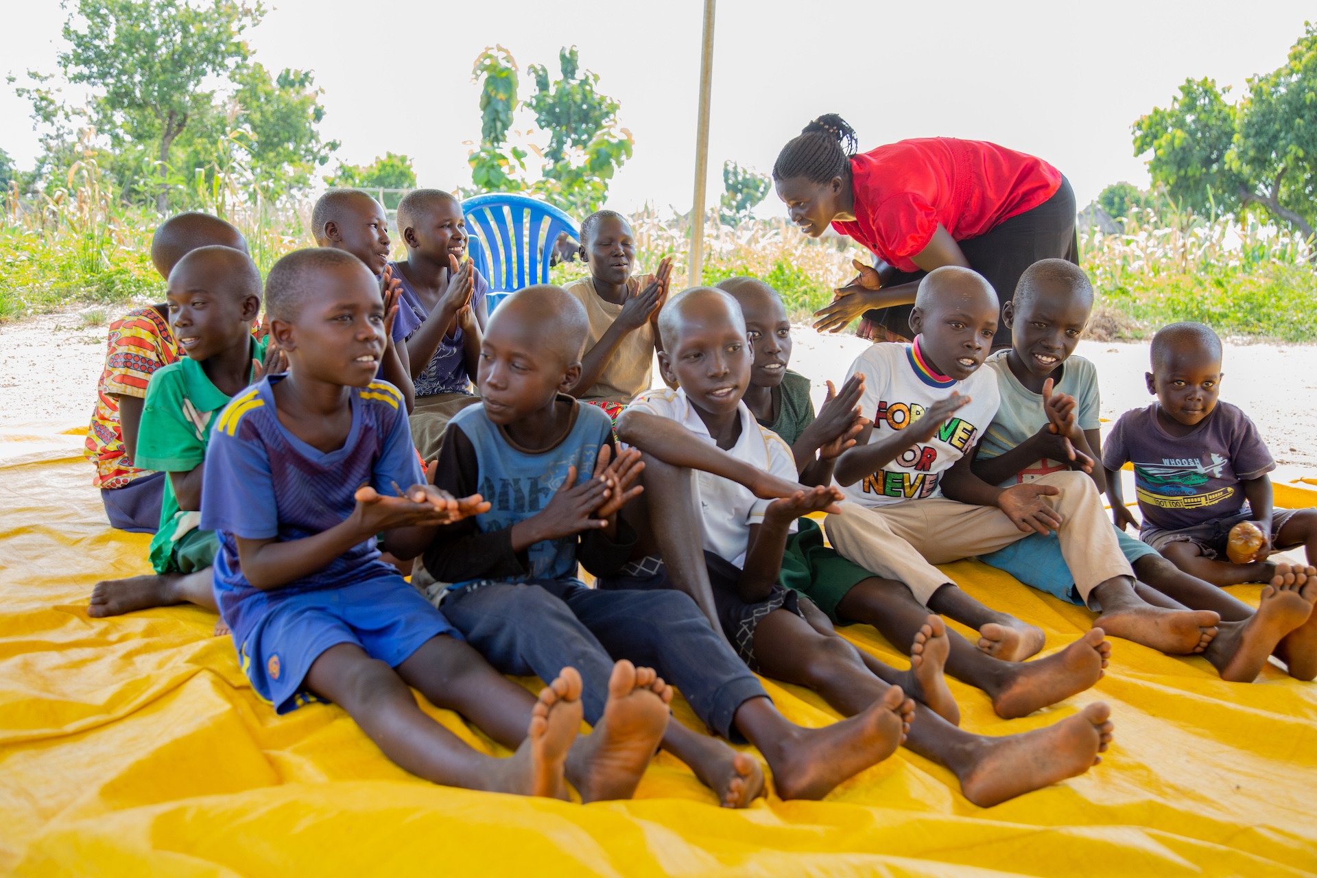 Children sit outside on a yellow tarpulin and play a hand clapping game while a teacher leans down to talk to one of them