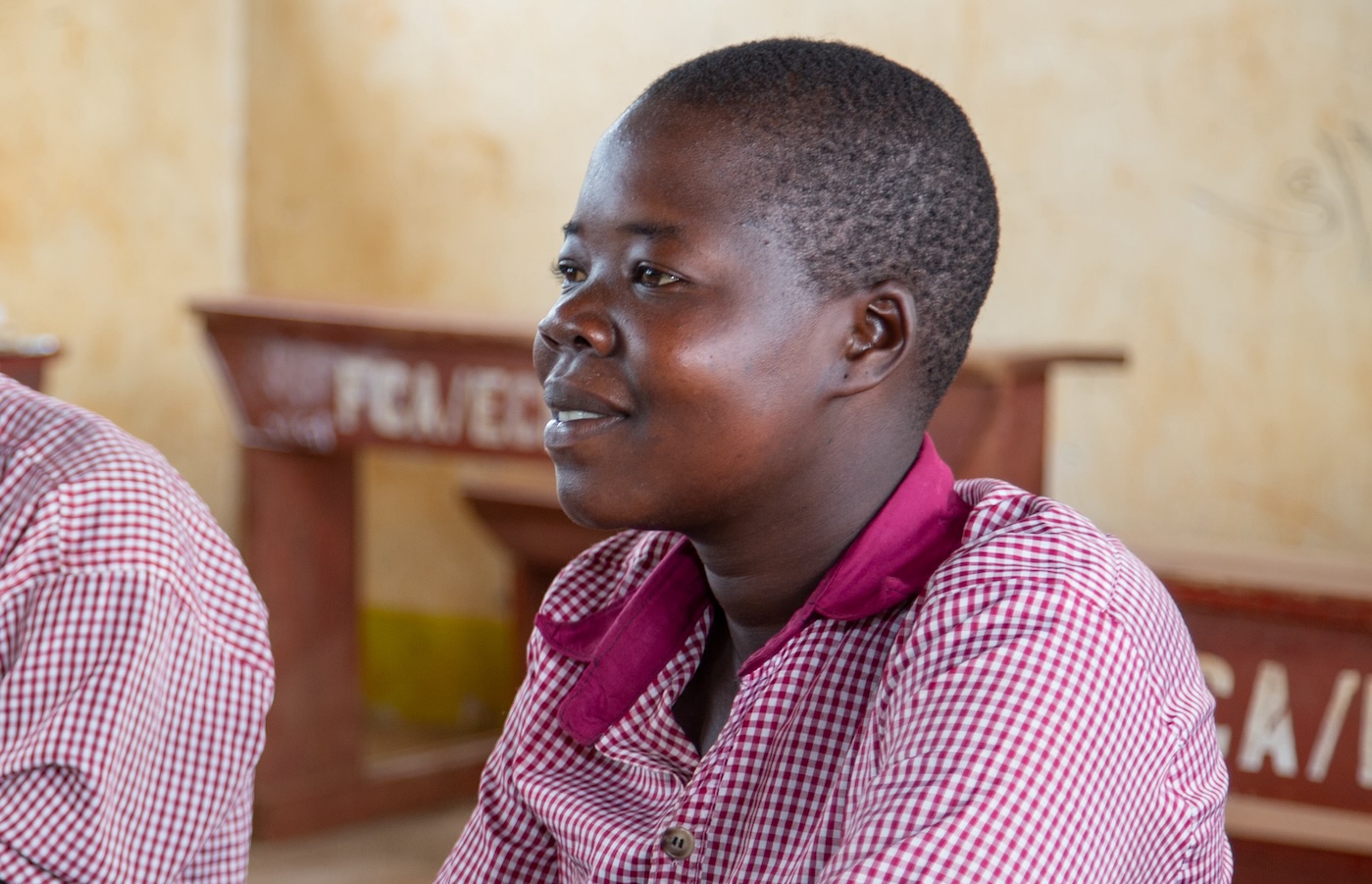 A teenager in school uniform sits at a desk in a classroom. She is listening and smiling.