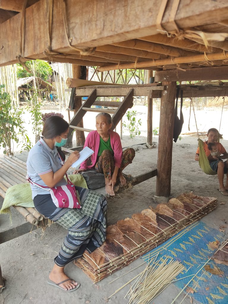 A woman in a face mask takes notes while sitting next to an elderly woman. They are sitting underneath a wooden hut. In the background, a man cradles a baby in a swing