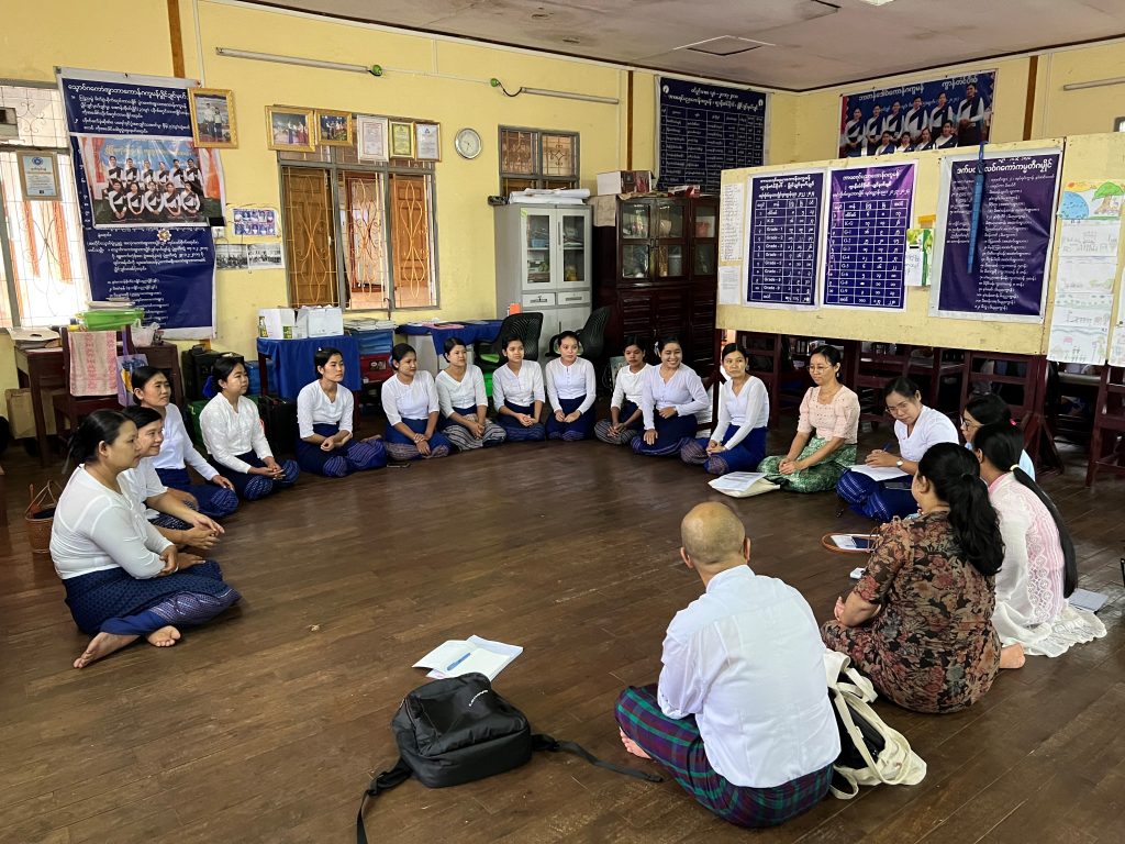 A group of adults sit in a circle on the floor in a classroom-like room, surrounded by posters on walls and boards