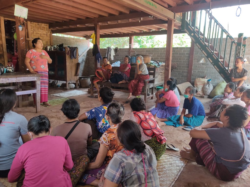 A woman stands talking to a large group of women sitting on the floor