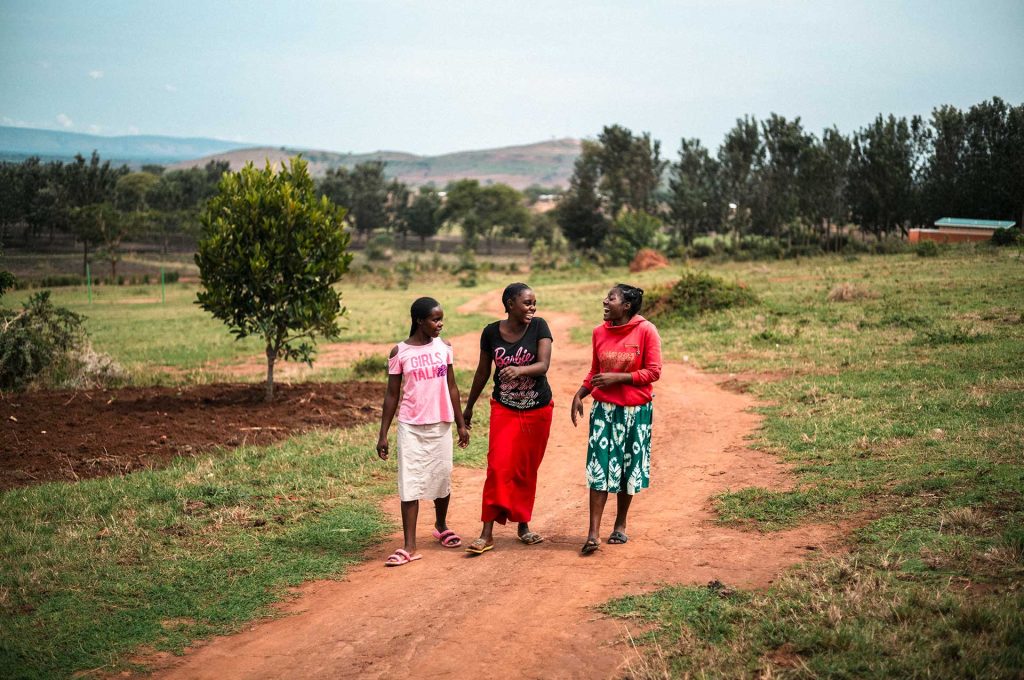 Tre unga kvinnor går på en sandväg i flyktingbosättningsområdet i Uganda. De skämtar och skrattar.