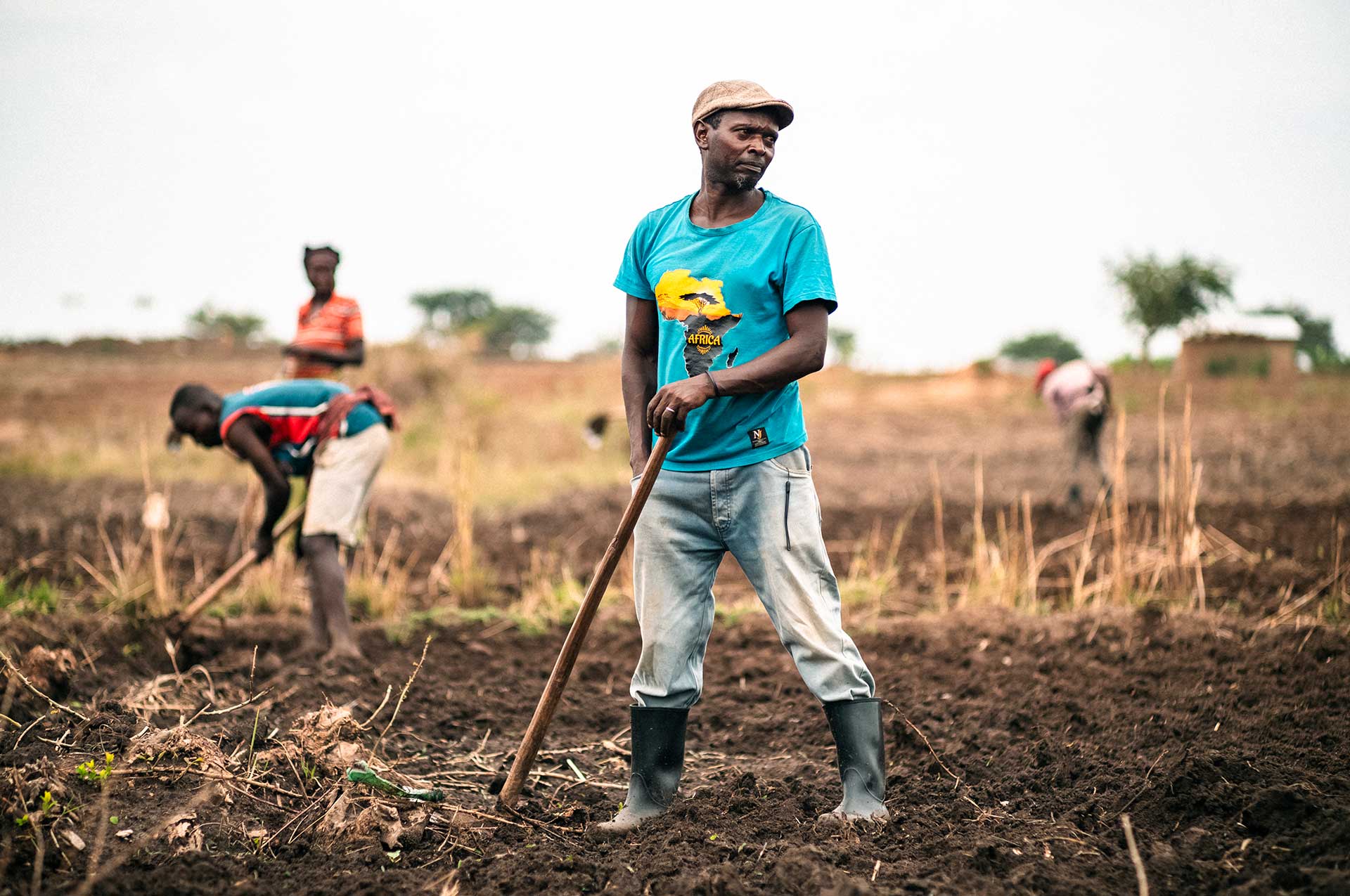 Lakkipäinen mies seisoo pellolla Ugandassa ja nojaa kuokkaansa. Taustalla näkyy maisema ja muita ihmisiä, jotka työskentelevät pellolla.