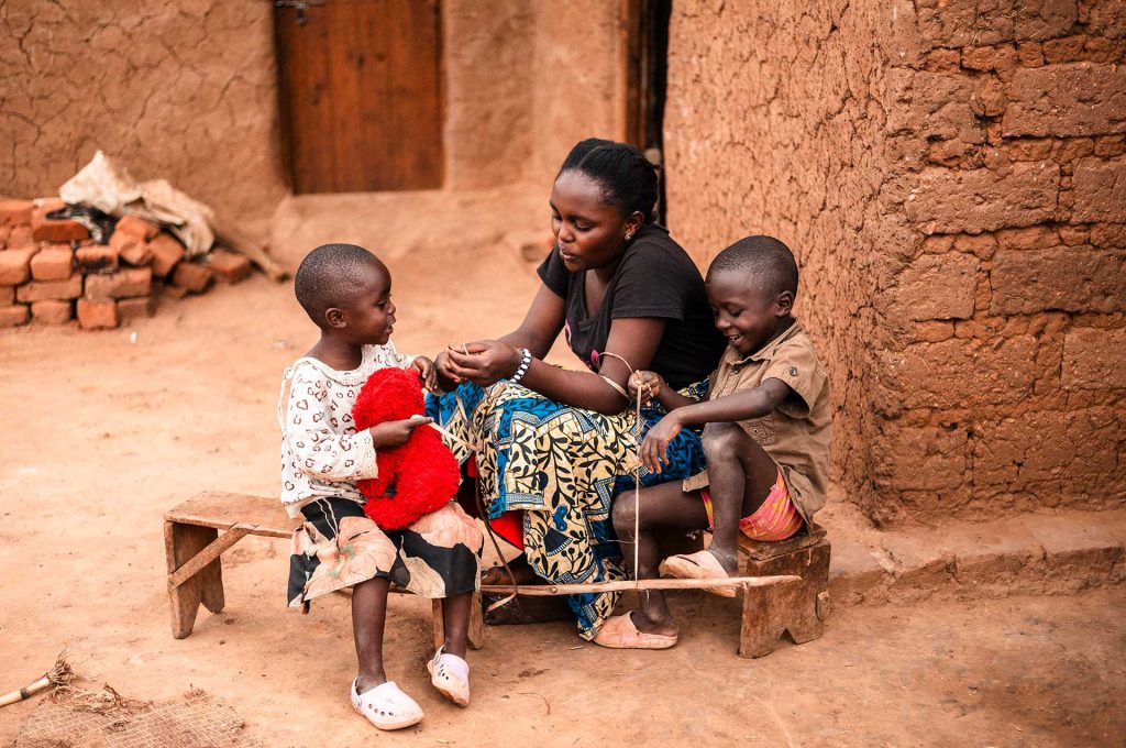 En ung kvinna sitter på en låg träbänk framför ett brunt tegelhus i Uganda och talar med två små barn. Ett av barnen har en teddybjörn i famnen. I bakgrunden syns en byggnad och en hög med tegelstenar.