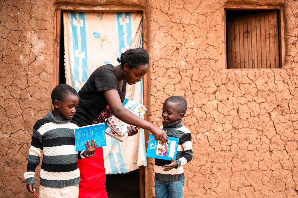 En ung kongolesisk kvinna ger ett häfte till ett barn som står framför henne. Framför kvinnan håller ett annat barn i en lärobok. Bakom dem syns en murad byggnad.