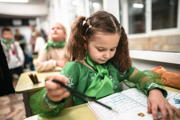 A young school girl is sitting by a window reading her notebook in a classroom. There are other little students seated behind the girl. 