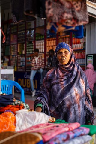 A woman in a patterned hijab and abaya stands behind a stall in a market