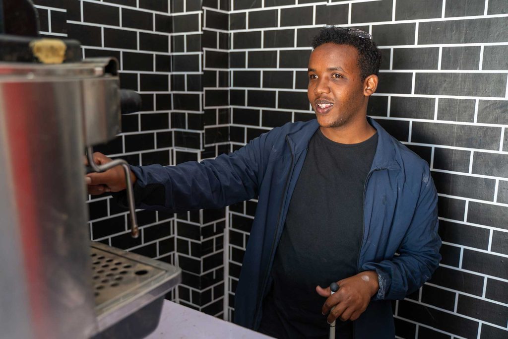 A man holding a white cane stands in front of a coffee making machine in front of a tiled wall. 