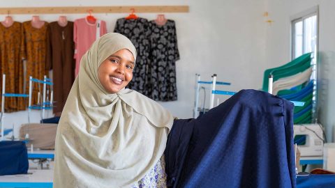 A woman presents the fabric she sewed in Somaliland. Behind the woman, you can see shirts hanging on hangers on the wall.