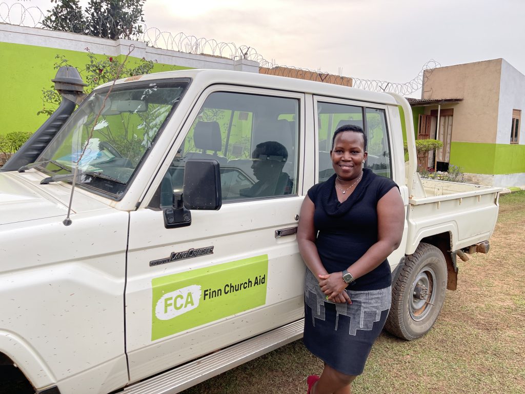 A smiling woman in an FCA-black T-shirt stands in front of a Land Cruiser bearing an FCA logo. 