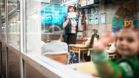 Kuva on otettu ukrainalaiseen luokkahuoneeseen ikkunan läpi. Luokkahuoneessa pieni tyttö vilkuttaa kameralle. Luokan edessä opettaja pitää oppituntia.