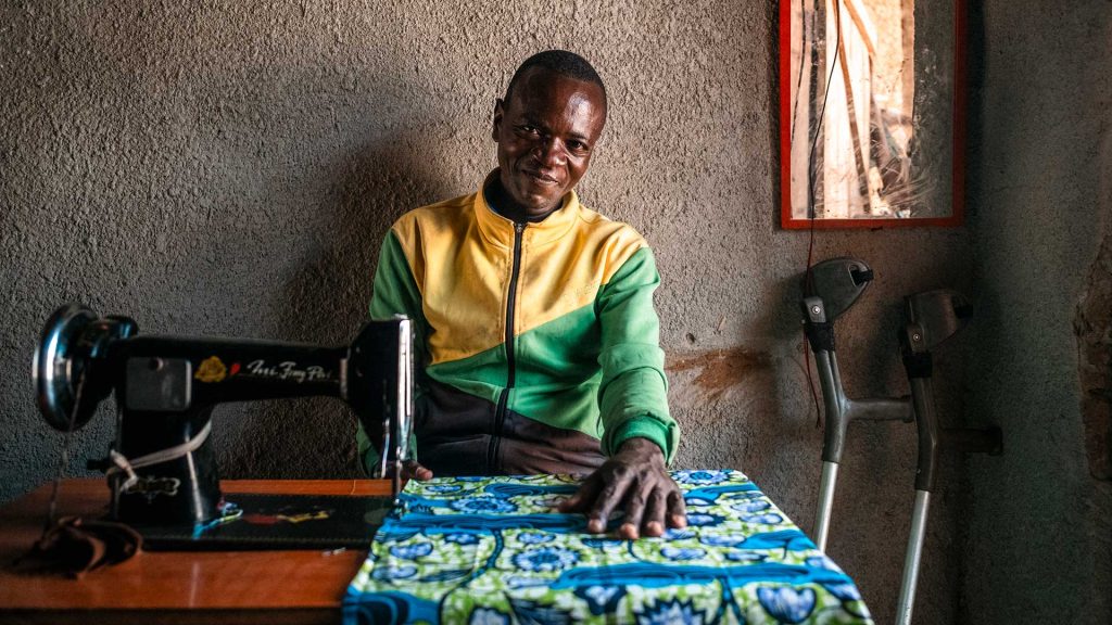 Mies istuu ompelemassa Ugandassa. Miehen edessä pöydällä on kangas, jota mies ompelee. Seinää vasten nojaavat kainalosauvat.