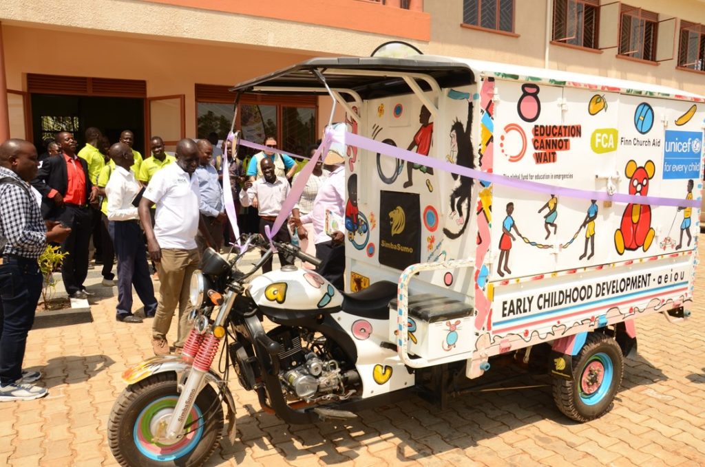 A three-wheeled motorcycle mounted with a trailer is standing outside. It has ribbons decorating it and the logos of Education Cannot Wait, FCA and UNICEF. People are smiling and inspecting it.