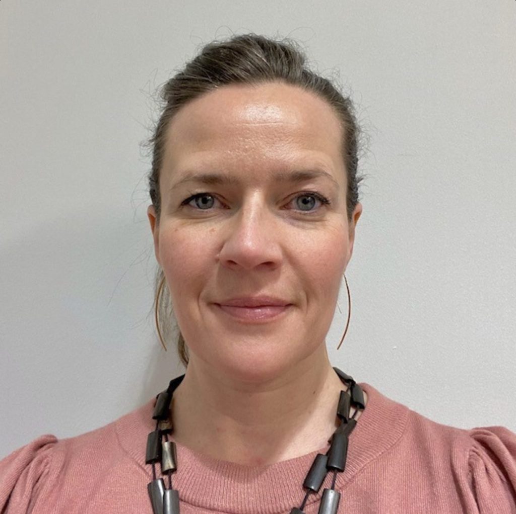 A woman in a pink top wearing wooden beads looks straight at the camera