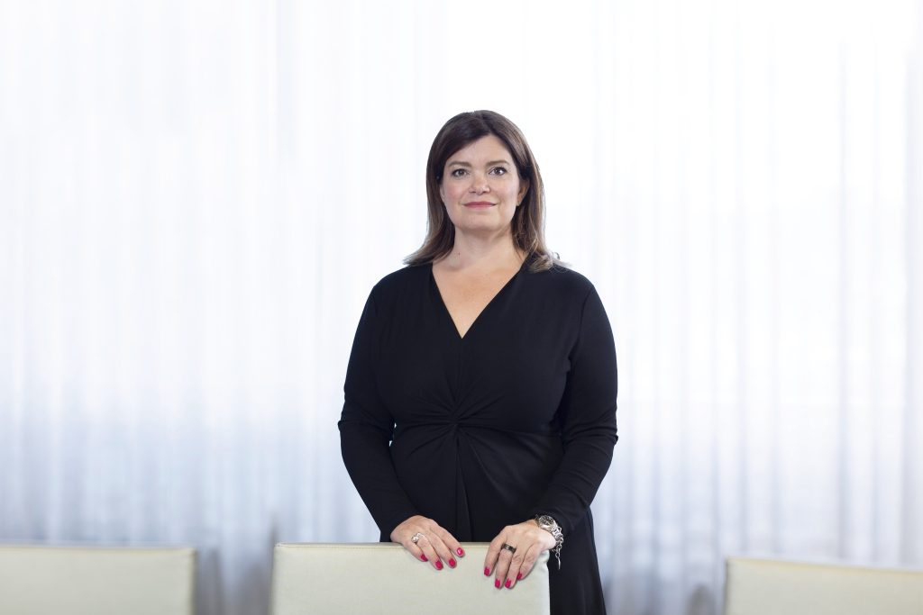 A woman in black stands behind a chair looking at the camera with a serene smile.