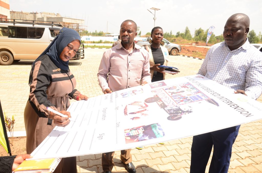 A woman signs a large cardboard document, which is held by two men. Another woman in the background is holding some files. They are all standing outside in a car park.