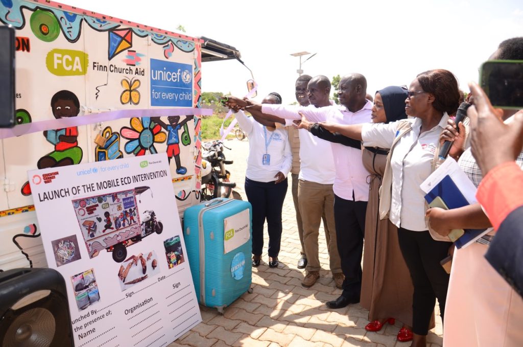 A line of men and women standing outside are smiling and pointing to a tuktuk, which has the logos of FCA and UNICEF on it, along with hand-drawn illustrations of children playing.