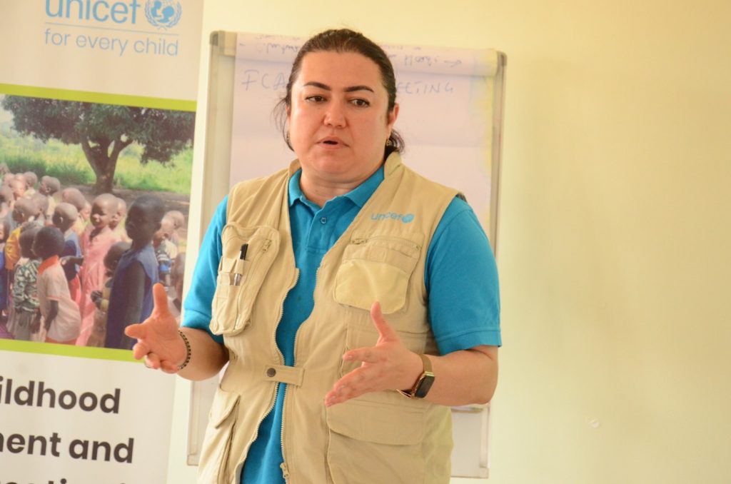 A woman in a blue shirt and a waistcoat with a UNICEF logo is talking in a room while standing in front of a flipchart and a banner which reads "UNICEF, for every child".