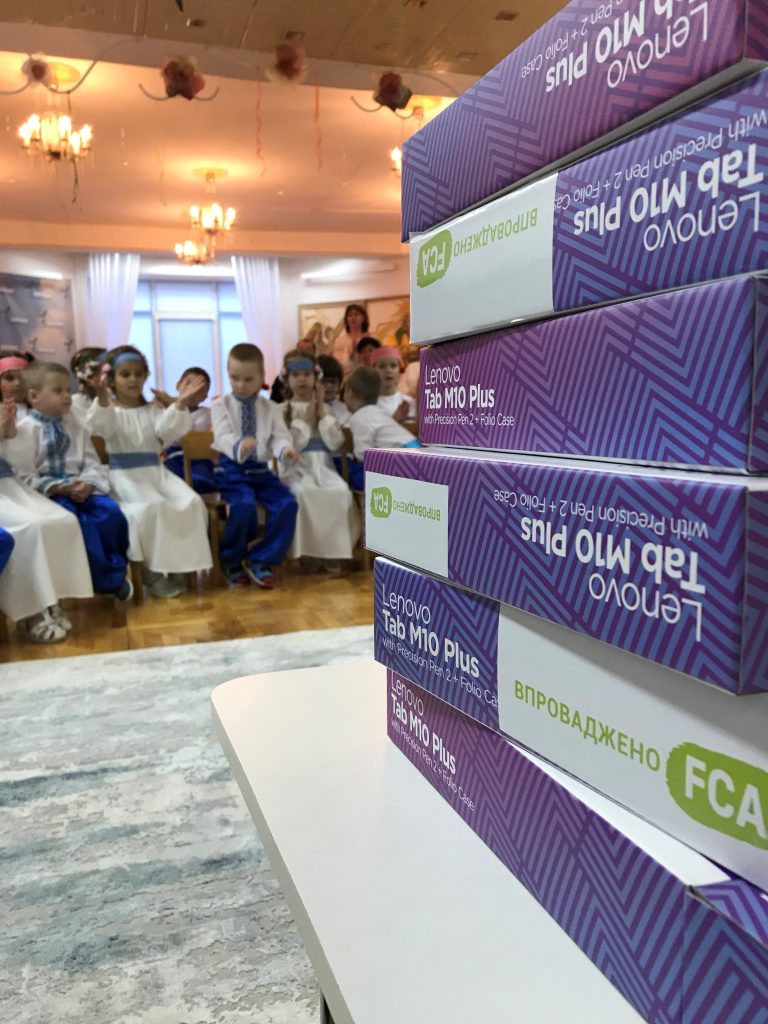 A pile of Lenovo tablets with the FCA logo sit in the foreground, while children sit in the background