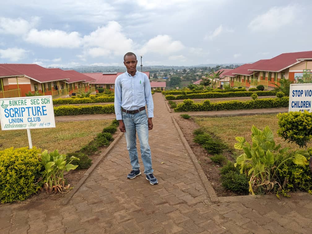 A young man stands on a path between neat border rows of plants. School buildings stand either side. 