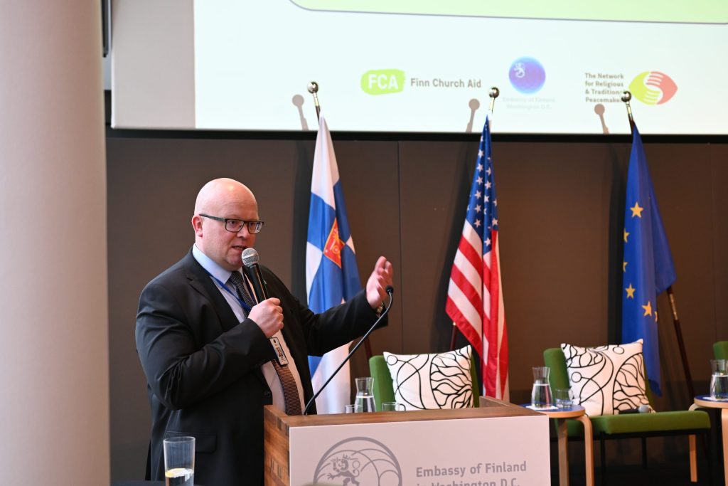 A man with a microphone speaks at a podium bearing the logo of the Embassy of Finland. 