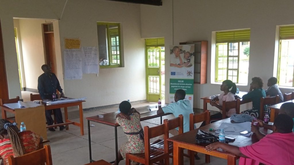 A number of adults sit in a classroom and listen to another teacher who stands at a desk at the front