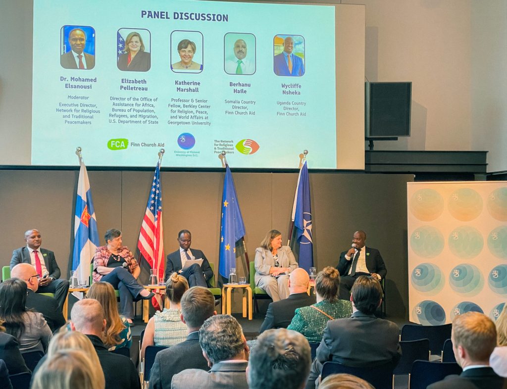 A panel of five people sit on a stage taking part in a discussion. The flags of Finland, the US, the EU and NATO are behind them. An audience sits in front.