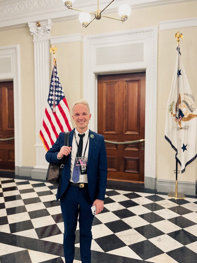 A smiling man in a suit carrying a rucksack poses for a photo in a hallway in front of a wooden door, which is flanked by the US flag and the flag of the Vice President of the United States.